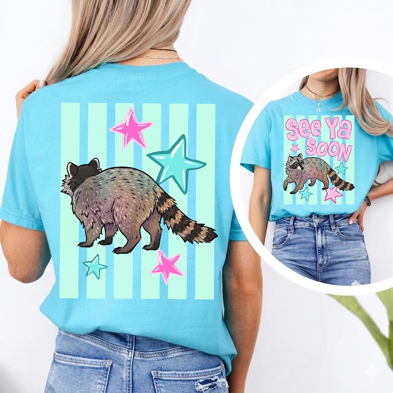 May include: Light blue t-shirt featuring a raccoon illustration with stars and vertical stripes. The raccoon is in shades of gray and brown. The shirt also has the text "See Ya Soon" on the front.