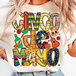 Puede incluir: Camiseta color crema con un gráfico colorido de "Cinco de Mayo". El diseño presenta elementos festivos como una guitarra, maracas, un sombrero y una botella de tequila, todo en colores y patrones vibrantes. El texto también está decorado con diseños florales y geométricos.
