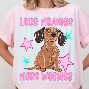 Könnte beinhalten: Rosafarbenes T-Shirt mit einer Cartoon-Dackel-Illustration. Das Shirt zeigt den Text "LESS MEANIES MORE WEENIES" in Rosa, mit Sternen und blauen Streifen im Hintergrund. Ein lustiges, lässiges T-Shirt.