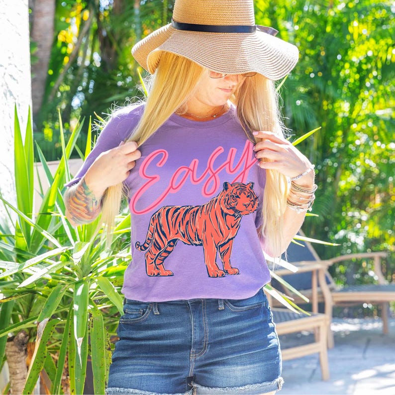 May include: A purple t-shirt with the word "Easy" in pink script and a red and orange tiger graphic. The shirt is paired with denim shorts and a straw hat.