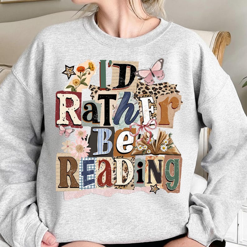 May include: A light gray sweatshirt with the text "I'D RATHER BE READING" in a collage style. The design includes floral and butterfly accents, with a mix of fonts and textures. The sweatshirt is a casual, comfortable style.