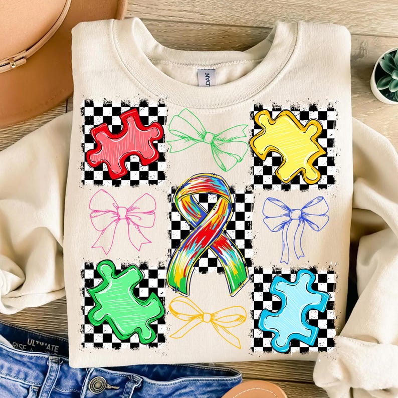 May include: Cream-coloured sweatshirt featuring colourful puzzle pieces, bows, and a rainbow ribbon design. The design is set against a black and white chequered background. The sweatshirt is a casual, comfortable style.