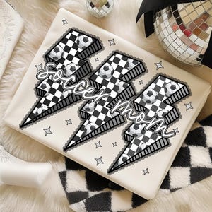 Puede incluir: Una manta color crema con tres dise&ntilde;os de rayos, cada uno con un patr&oacute;n de cuadros blancos y negros y balones de f&uacute;tbol. Las palabras "Soccer Mom" est&aacute;n escritas sobre los rayos. Peque&ntilde;as estrellas decoran la manta.