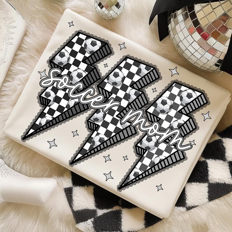 Puede incluir: Una manta color crema con tres dise&ntilde;os de rayos, cada uno con un patr&oacute;n de cuadros blancos y negros y balones de f&uacute;tbol. Las palabras "Soccer Mom" est&aacute;n escritas sobre los rayos. Peque&ntilde;as estrellas decoran la manta.