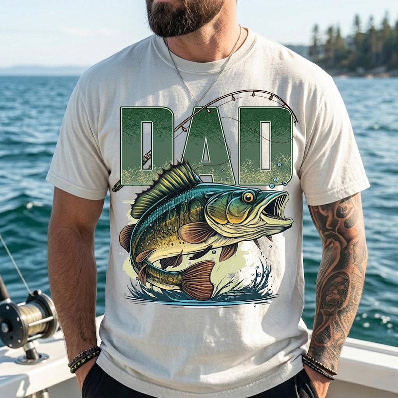 Pu&ograve; includere: T-shirt color crema con un'illustrazione dettagliata di un pesce che salta fuori dall'acqua. La parola "DAD" &egrave; stampata sopra il pesce in un carattere verde strutturato, con un disegno di canna da pesca. La maglietta &egrave; indossata da una persona su una barca.