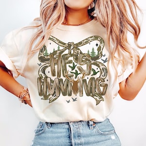 May include: Cream-colored t-shirt with the text "HE'S HUNTING" in a camouflage-style font. The design includes a bow, trees, birds, and a deer head. The shirt is made of a soft material and is suitable for casual wear.