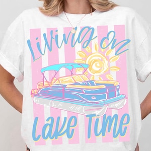 May include: White t-shirt with a graphic of a blue and white pontoon boat, a yellow sun, and the text "Living on Lake Time" in blue script. The background has pink and white vertical stripes.