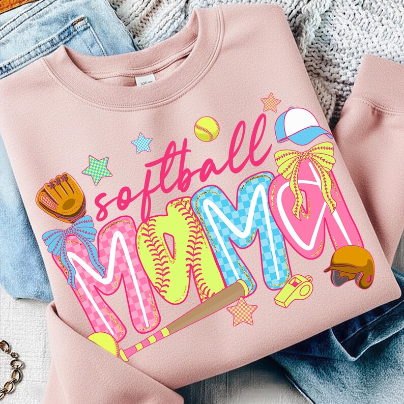 May include: A light pink sweatshirt with the words "softball MAMA" in colorful, playful lettering. The design includes a baseball glove, stars, a baseball, a hat, a whistle, and a helmet, all in a cartoon style.