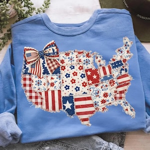 Peut inclure: Sweat-shirt bleu clair orné d'un motif patchwork représentant la carte des États-Unis. La carte est composée de divers motifs de tissu rouges, blancs et bleus, avec des étoiles, des rayures et des imprimés floraux. Un nœud avec un motif de drapeau américain est en haut.