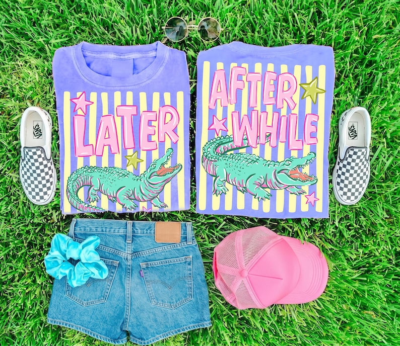 May include: Two purple t-shirts with the phrases "LATER" and "AFTER WHILE" and a cartoon alligator graphic. The image also includes denim shorts, a pink trucker hat, a blue scrunchie, checkered slip-on sneakers, and sunglasses, all arranged on green grass.