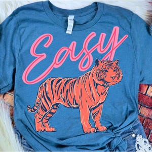 May include: Blue t-shirt with the word "Easy" in pink script above an orange and navy tiger illustration. The tiger is standing, facing forward, with detailed stripes and facial features. The shirt is a casual style.