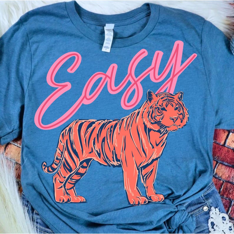May include: Blue t-shirt with the word "Easy" in pink script above an orange and navy tiger illustration. The tiger is standing, facing forward, with detailed stripes and facial features. The shirt is a casual style.