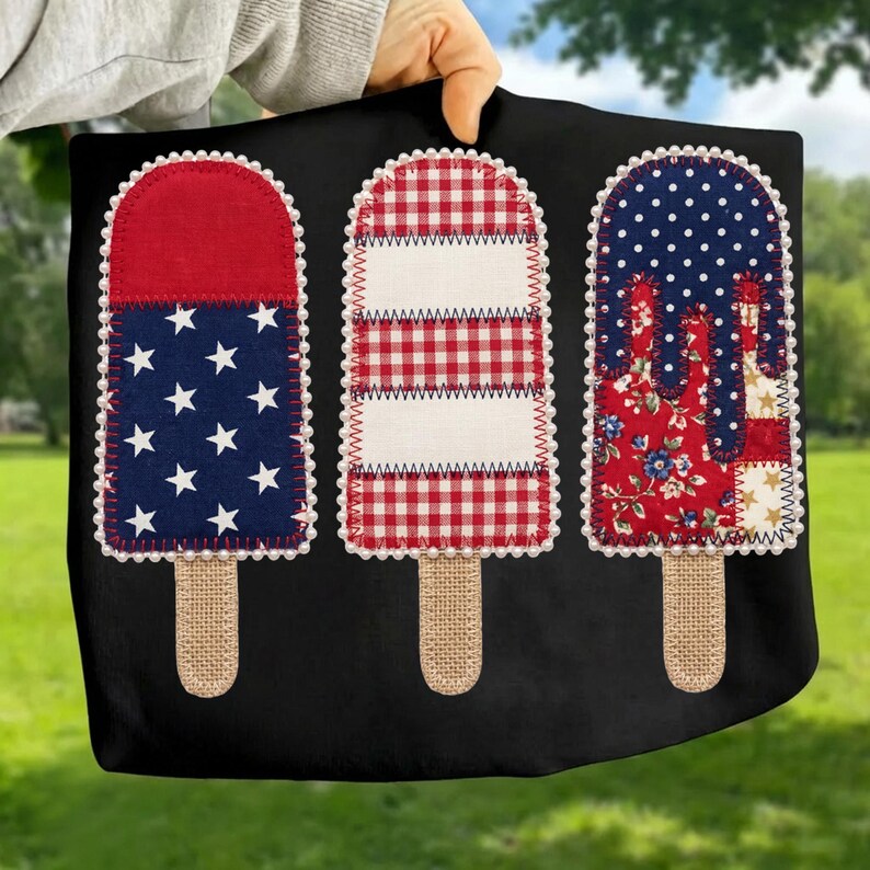 May include: Black tote bag with three decorative popsicle designs. Each popsicle has a different patriotic pattern: red and white stripes, blue with white stars, and a floral pattern. The popsicles have burlap stick handles and are outlined with white beads.