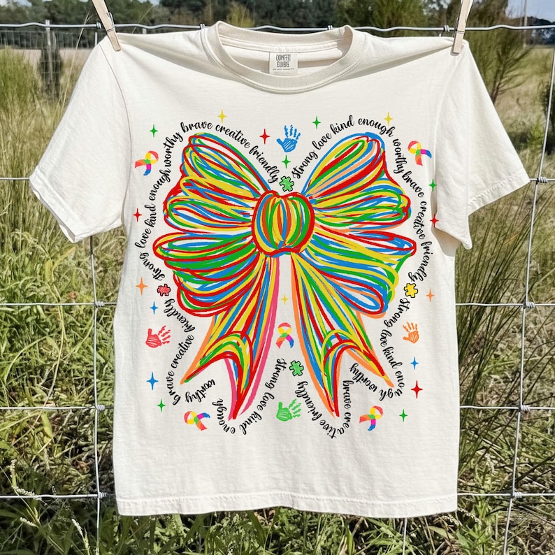 May include: Cream-colored t-shirt featuring a large, colorful bow design with the words "brave, creative, friendly, strong, love, kind, enough, worthy" around the bow. The shirt is hanging on a clothesline.