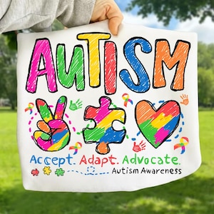May include: White fabric with the word "AUTISM" in colorful, hand-drawn letters. Below are a peace sign, puzzle piece, and heart, all in rainbow colors, with the words "Accept. Adapt. Advocate. Autism Awareness."