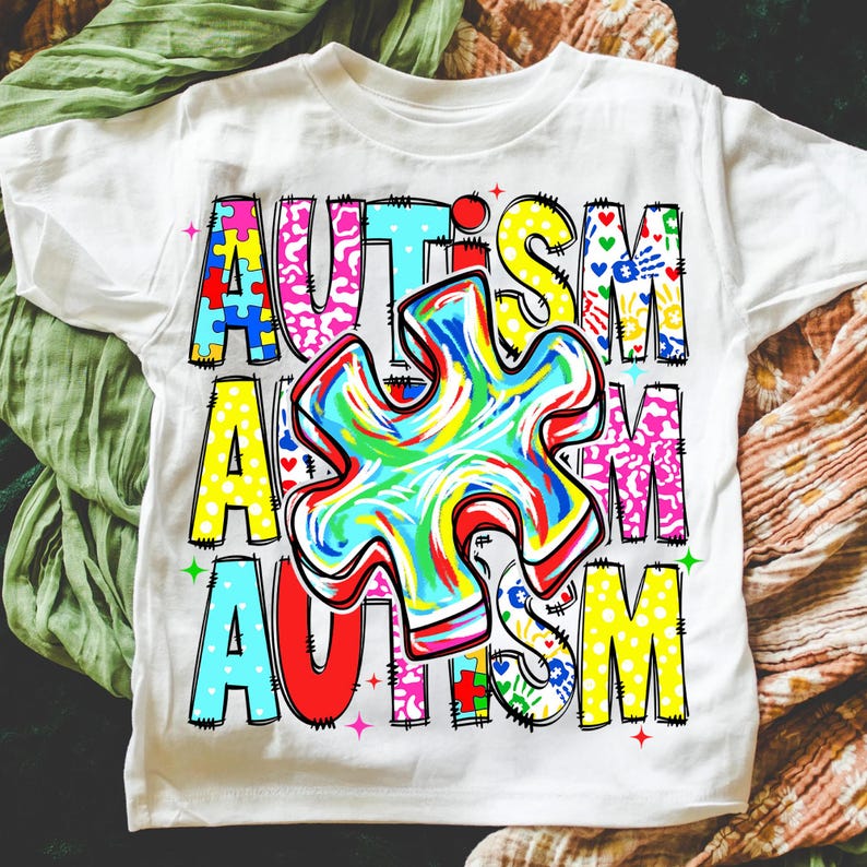 May include: White t-shirt with the word "AUTISM" in colorful, block letters. The letters are filled with puzzle pieces, hearts, and other designs. A large, multi-colored puzzle piece is in the center of the shirt.