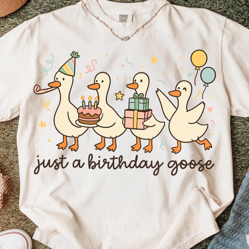 May include: Cream-colored t-shirt featuring a cartoon illustration of four geese celebrating a birthday. The geese are carrying a cake, gifts, and balloons. The text "just a birthday goose" is printed below the illustration.