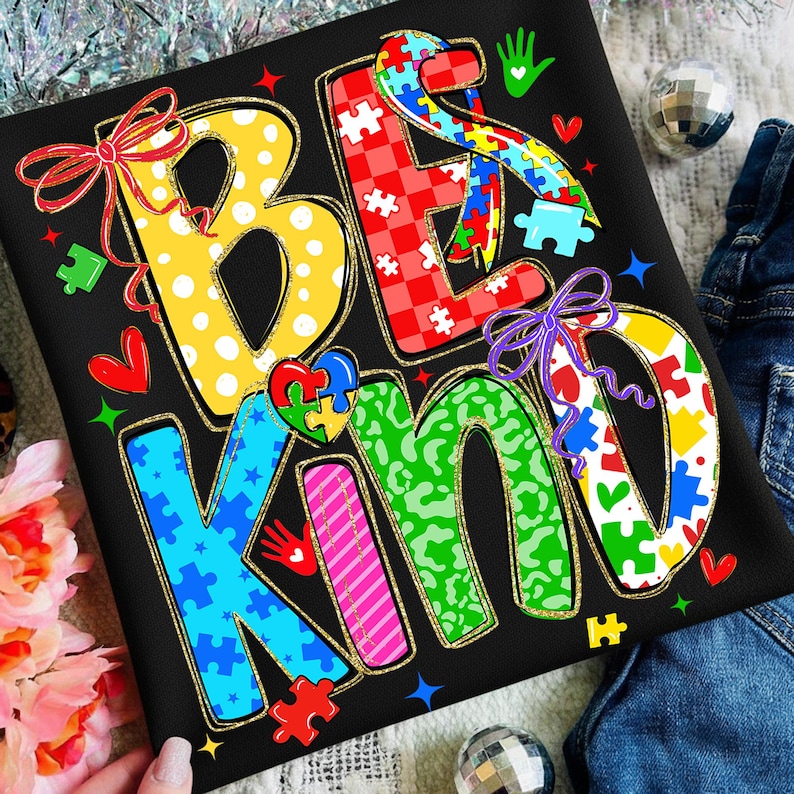 May include: Black t-shirt with the words "BE KIND" in colorful, puzzle-piece-themed letters. The letters are outlined in gold and decorated with bows, hearts, and stars. The design has an autism awareness theme.
