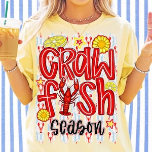 May include: Yellow t-shirt with the words "CRAWFISH SEASON" in red outlined letters. The design includes a red crawfish, lemon slices, stars, and a sunflower. The background has a pattern of crawfish and blue and white stripes.