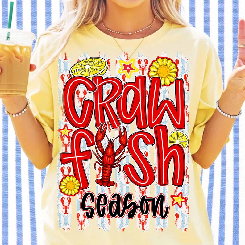 May include: Yellow t-shirt with the words "CRAWFISH SEASON" in red outlined letters. The design includes a red crawfish, lemon slices, stars, and a sunflower. The background has a pattern of crawfish and blue and white stripes.