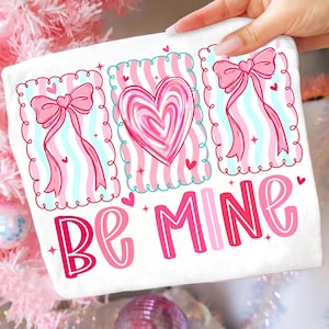 May include: White Valentine's Day themed decorative pillow with pink and red accents. The pillow features three framed designs with a heart and bows, and the words "Be Mine" in a playful font.