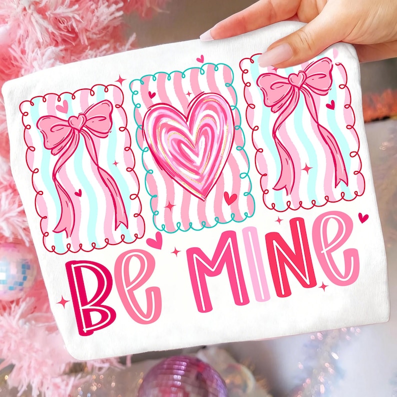 May include: White Valentine's Day themed decorative pillow with pink and red accents. The pillow features three framed designs with a heart and bows, and the words "Be Mine" in a playful font.