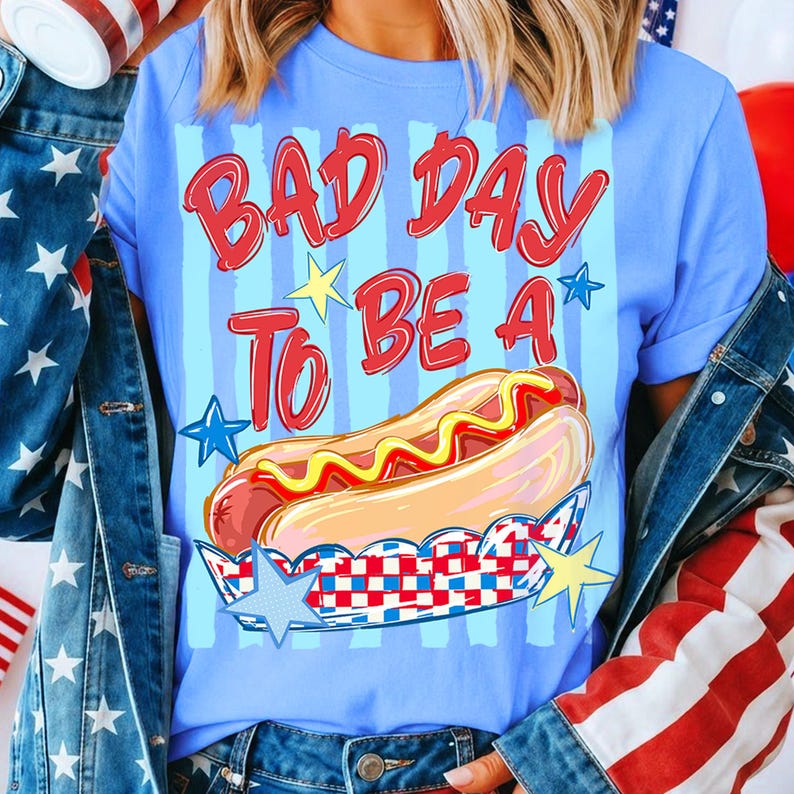 以下が含まれることがあります： 赤色の「BAD DAY TO BE A」の文字とホットドッグのイラストが描かれた水色のTシャツ。 ホットドッグは赤、白、青の容器に入っており、黄色と青の星が描かれています。 このTシャツはカジュアルなスタイルです。