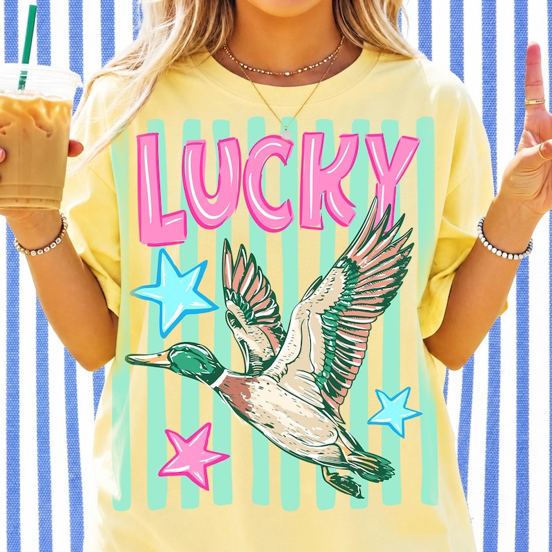 Puede incluir: Camiseta amarilla con la palabra "LUCKY" en rosa. Debajo, un gr&aacute;fico de un pato volando con detalles verdes, rosas y marrones, y estrellas azules y rosas. La camiseta tiene un corte holgado.