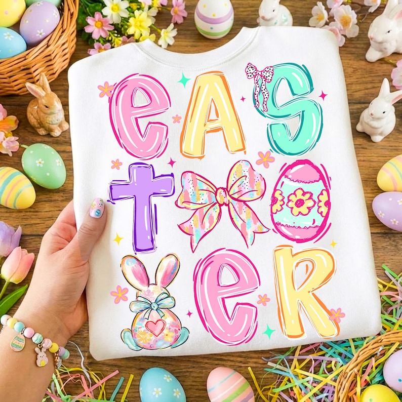 May include: White Easter sweatshirt with colorful text and graphics. The word "EASTER" is displayed in bubble letters with a cross, bow, egg, and bunny illustrations. Decorative Easter eggs and bunnies surround the sweatshirt.