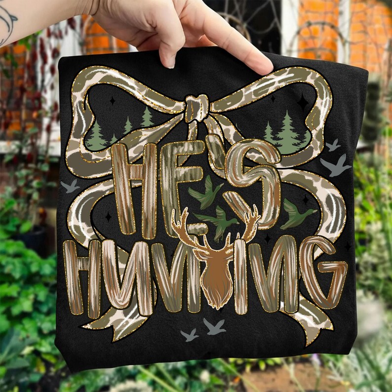 May include: Black t-shirt with the text "HE'S HUNTING" in a camouflage and wood-grain design. The design includes a bow, deer, trees, and flying birds. The shirt is held up by a hand.