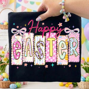 Może przedstawiać: Czarny t-shirt z napisem "Happy Easter" w kolorowych, dekoracyjnych literach. Litery zdobione są różnymi wzorami i kolorami, w tym różowym, żółtym i niebieskim, z kwiatowymi i kokardkowymi akcentami. Koszulka trzymana jest przez dłoń.
