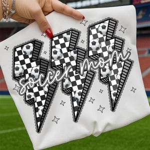 Puede incluir: Camiseta blanca con un dise&ntilde;o gr&aacute;fico. El dise&ntilde;o presenta las palabras "Soccer Mom" en una fuente de escritura, balones de f&uacute;tbol y un patr&oacute;n de cuadros blanco y negro dentro de formas de rayo, delineado con un ribete de encaje.