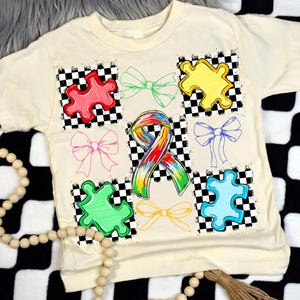 May include: Cream-coloured t-shirt featuring colourful puzzle pieces and bows on a chequered background. A rainbow ribbon is centered on the shirt. The shirt is a casual style.