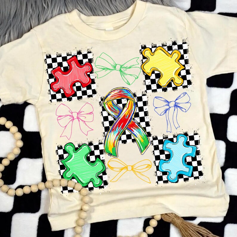 May include: Cream-coloured t-shirt featuring colourful puzzle pieces and bows on a chequered background. A rainbow ribbon is centered on the shirt. The shirt is a casual style.