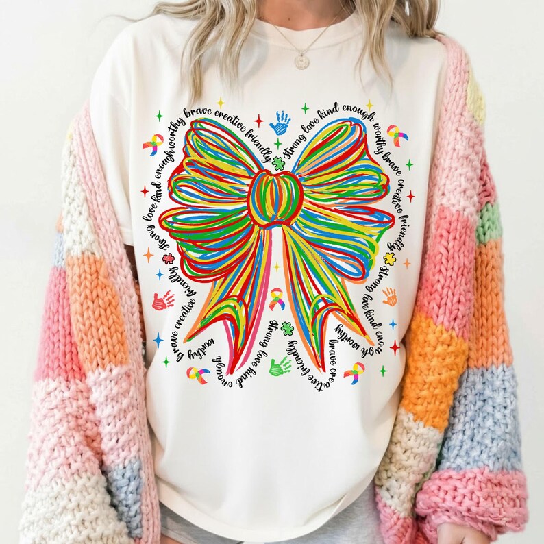 May include: Cream-colored t-shirt featuring a large, colorful bow design with the words "brave, creative, friendly, strong, love, kind, enough, worthy" around the bow. The shirt is paired with a multi-colored, striped cardigan.