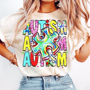May include: A cream-colored t-shirt with the word "AUTISM" in colorful, block letters. The letter "A" is in blue, the "U" is in pink, the "T" is in turquoise, the "I" is in yellow, the "S" is in pink, and the "M" is in yellow. A colorful puzzle piece is in the center.
