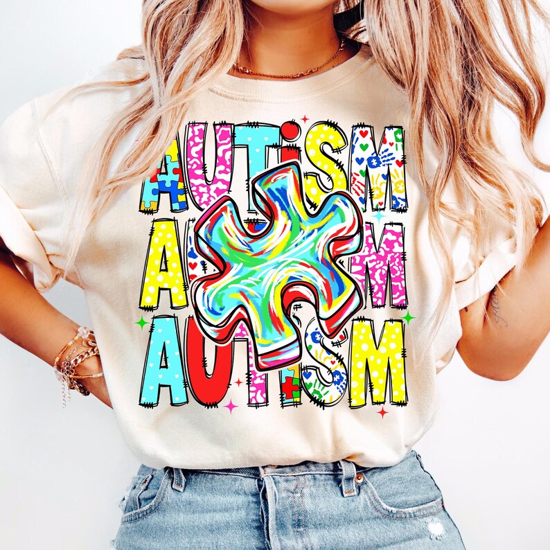 May include: A cream-colored t-shirt with the word "AUTISM" in colorful, block letters. The letter "A" is in blue, the "U" is in pink, the "T" is in turquoise, the "I" is in yellow, the "S" is in pink, and the "M" is in yellow. A colorful puzzle piece is in the center.