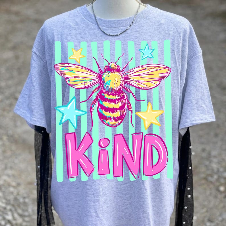 Puede incluir: Camiseta gris con un dise&ntilde;o de abeja colorida, alas amarillas y cuerpo rosa y amarillo. La palabra "KIND" est&aacute; impresa en rosa debajo, con estrellas azules y amarillas y rayas verdes.