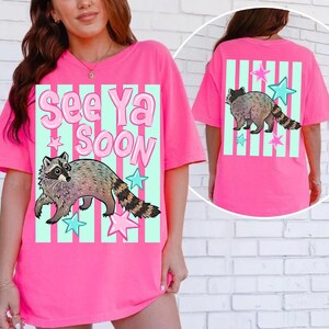 May include: Bright pink t-shirt featuring a raccoon graphic with the text "See Ya Soon". The design includes a raccoon, stars, and vertical stripes in shades of blue and pink. The back of the shirt also has a raccoon graphic.