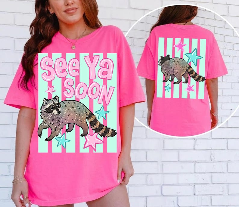 May include: Bright pink t-shirt featuring a raccoon graphic with the text "See Ya Soon". The design includes a raccoon, stars, and vertical stripes in shades of blue and pink. The back of the shirt also has a raccoon graphic.