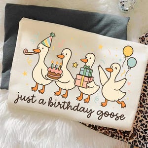 May include: Cream-colored graphic tee featuring a line of cartoon geese celebrating a birthday. One goose wears a party hat and carries a cake, others hold gifts and balloons. The text "just a birthday goose" is written below.