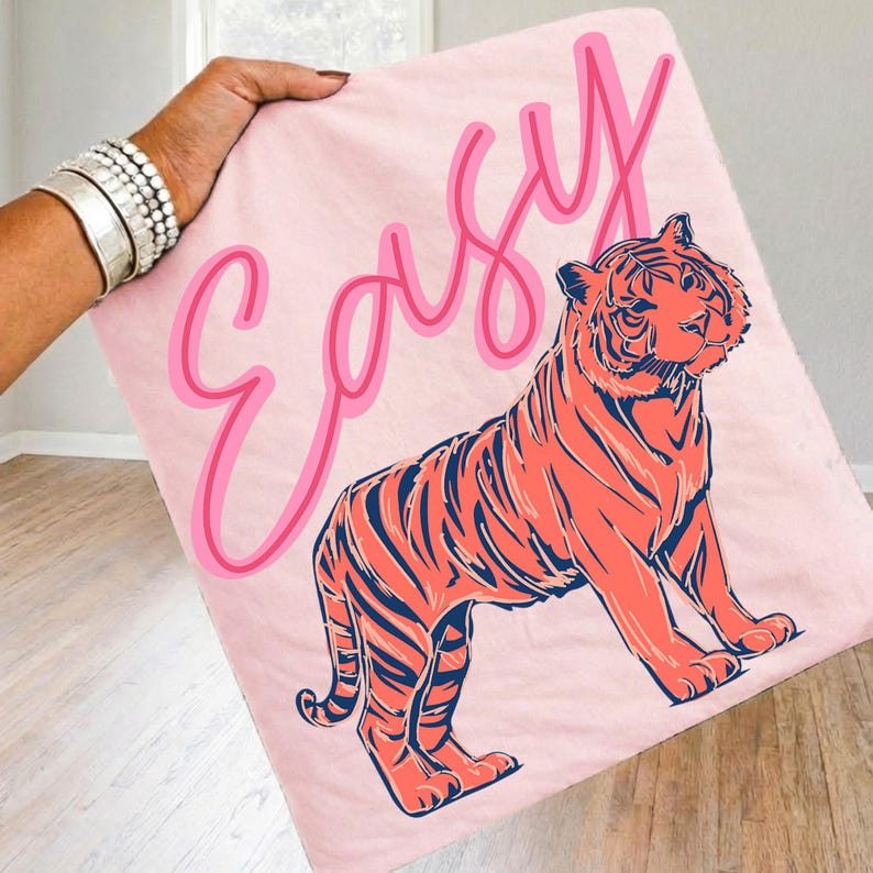 May include: A light pink blanket featuring a stylized orange tiger with navy blue stripes. The word "Easy" is written in pink script above the tiger. The blanket is held up by a person wearing silver bracelets.