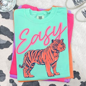 May include: A stack of folded t-shirts in shades of pink, orange, and teal. The teal shirt features the word "Easy" in pink script above a tiger illustration in orange and navy blue. The shirts are on a white and black patterned surface.