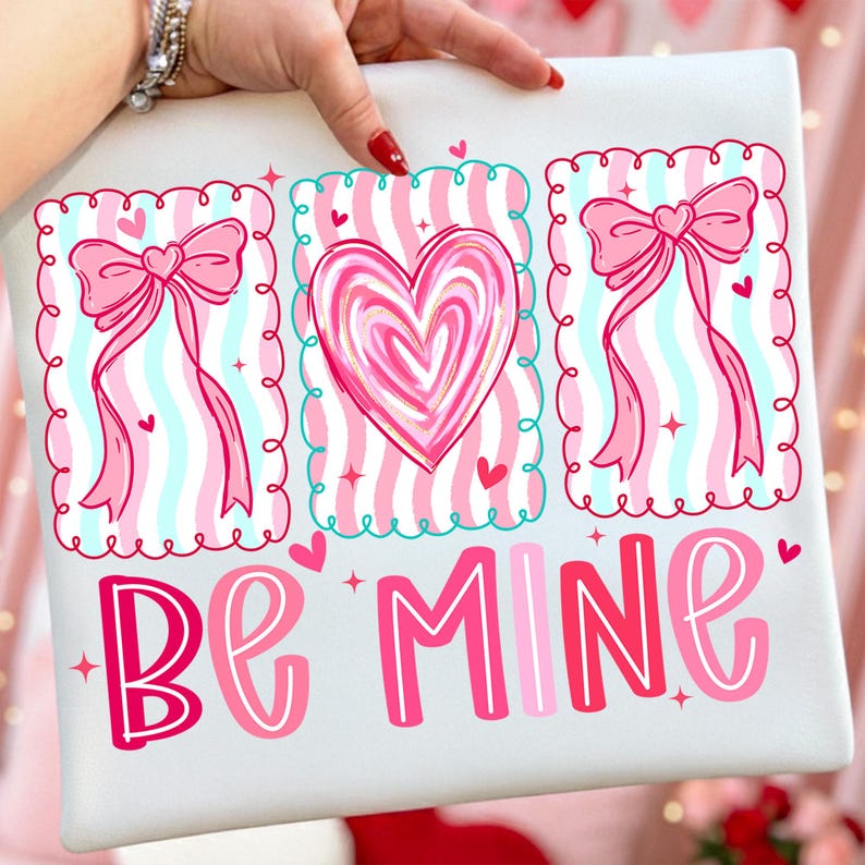 May include: A white pillow with a Valentine's Day design. The pillow features three framed illustrations with pink bows, hearts, and striped backgrounds. The words "Be Mine" are written in pink and red script across the bottom.