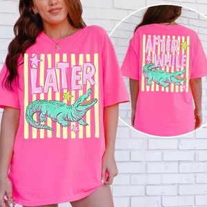 May include: A bright pink t-shirt with a graphic of a teal alligator and the word "LATER" in pink. The back of the shirt has the words "AFTER WHILE" and another alligator graphic. Yellow stripes and star accents complete the design.
