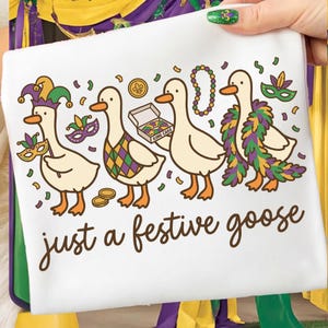 Op de afbeelding: Witte decoratieve handdoek met cartoon ganzen in feestelijke kleding. De ganzen zijn versierd met Mardi Gras-accessoires, waaronder maskers, kralen en een gevederde boa. De tekst "just a festive goose" is geschreven in een scriptlettertype.