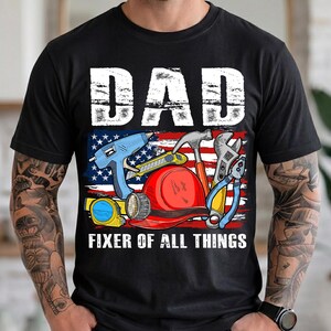 May include: Black t-shirt with a graphic featuring the word "DAD" in white block letters above an American flag design. The design includes tools like a hammer, wrench, and hard hat, with the text "FIXER OF ALL THINGS" below.