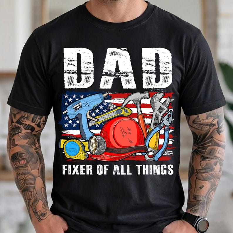 May include: Black t-shirt with a graphic featuring the word "DAD" in white block letters above an American flag design. The design includes tools like a hammer, wrench, and hard hat, with the text "FIXER OF ALL THINGS" below.