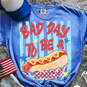 以下が含まれることがあります： 「BAD DAY TO BE A」の赤い文字とマスタードのかかったホットドッグ2つのグラフィックがプリントされた青いTシャツ。星と赤、白、青の配色が特徴です。背景には青と白の野球帽が見えます。