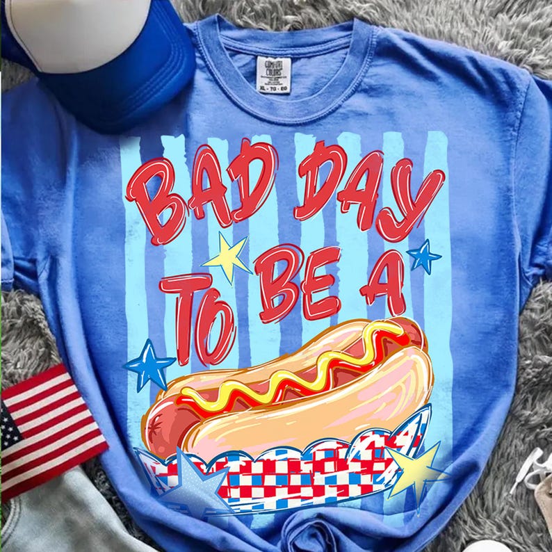 以下が含まれることがあります： 「BAD DAY TO BE A」の赤い文字とマスタードのかかったホットドッグ2つのグラフィックがプリントされた青いTシャツ。星と赤、白、青の配色が特徴です。背景には青と白の野球帽が見えます。